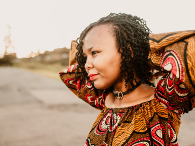 Christine stands with her arms behind her head, wearing an African-style dress and looking into the distance