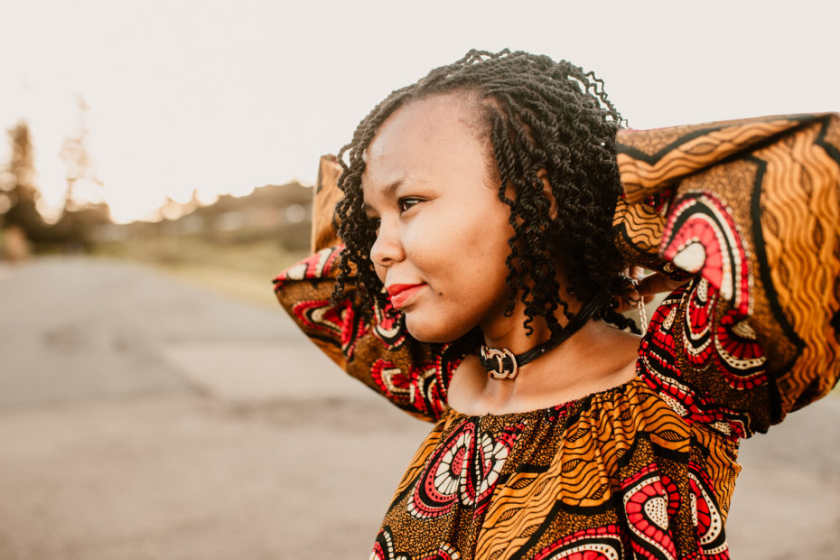 Christine stands with her arms behind her head, wearing an African-style dress and looking into the distance