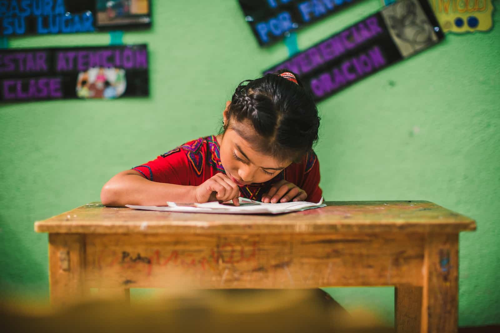 A girl from Guatemala in a red shirt with her hair pulled back sits at a wooden school desk, reading a letter. She is in front of a green wall. 