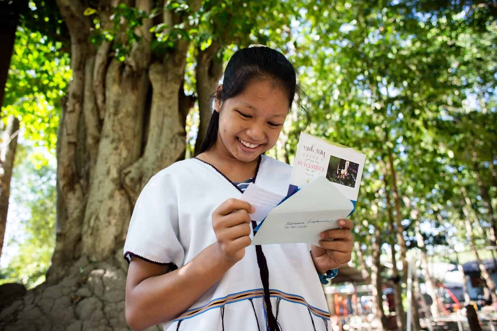 A girl from Thailand wearing a white traditional Karen tribe shirt smile while reading a card that she holds in her hand.