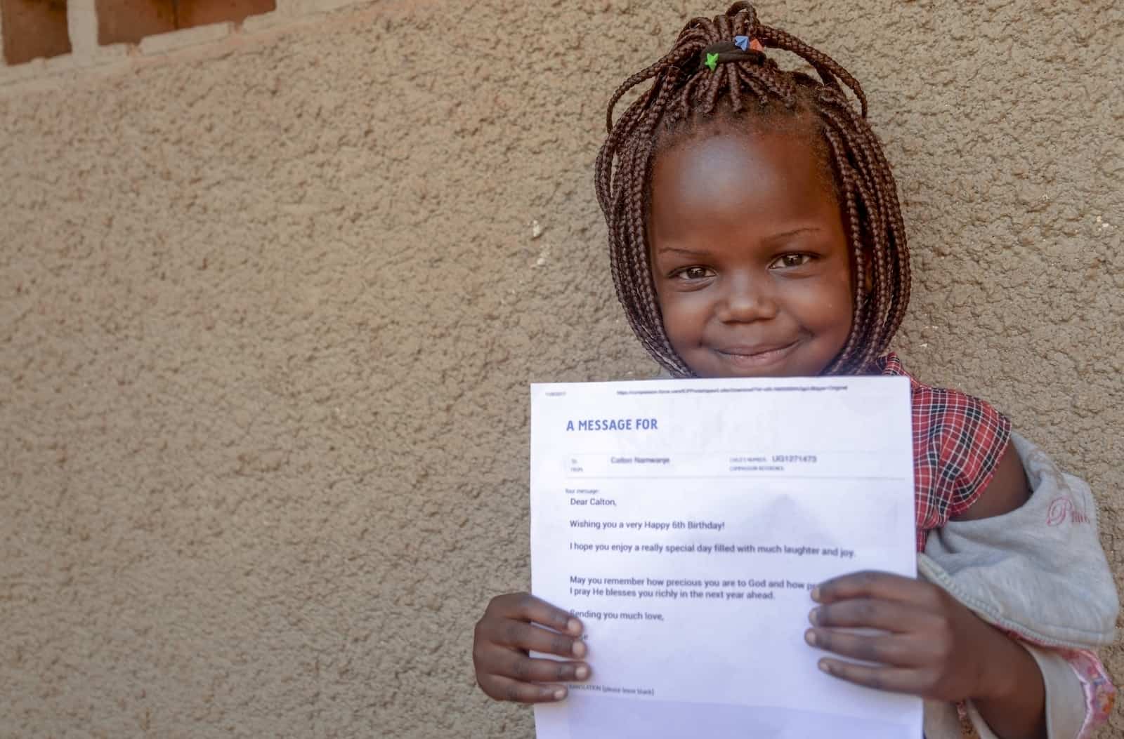 A young girl from Uganda with her hair in braids and a checkered shirt holds a letter in front of her and smiles.