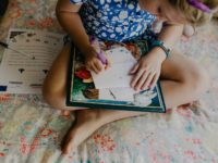 A girl with blonde hair and a blue dress on sits on her bed, legs crossed, writing a letter to the child she sponsors.