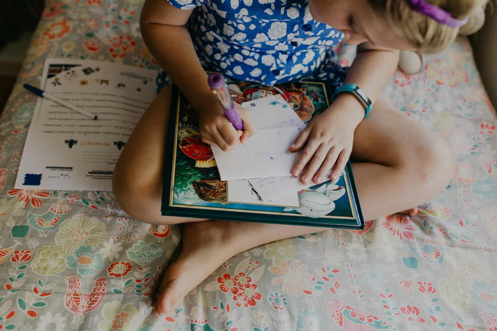 A girl with blonde hair and a blue dress on sits on her bed, legs crossed, writing a letter to the child she sponsors.