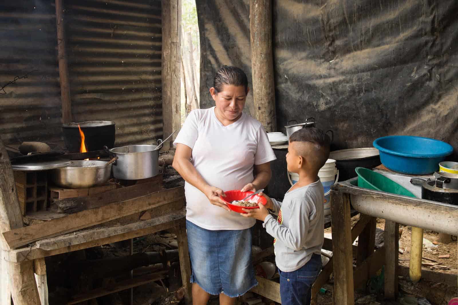 A woman stands in a kitchen handing a bowl of food to a boy.