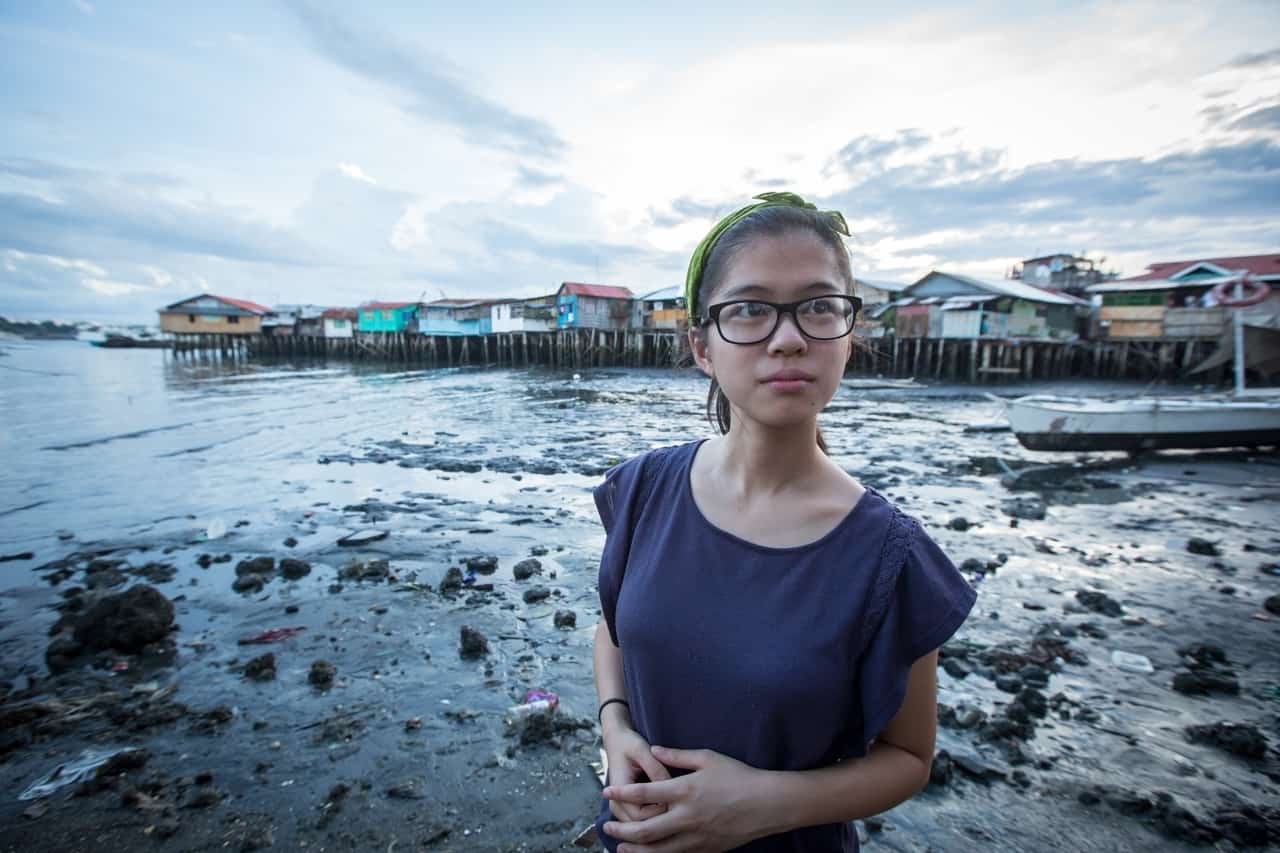 A girl wearing glasses and a blue shirt clasps her hands in front of her and looks to the side, standing in front of a waterway with homes on stilts in the background.