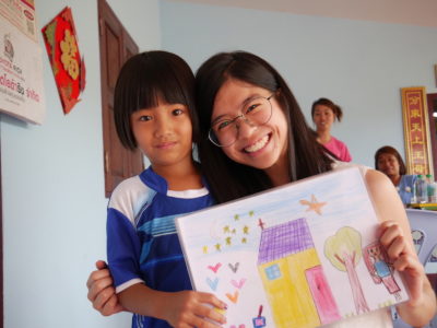 A young woman with glasses on puts her arm around a girl holding a drawing of a house.