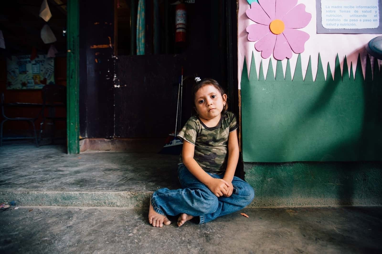 A little girl wearing a camouflage shirt, jeans and bare feet, sits on a concrete step, looking sad.