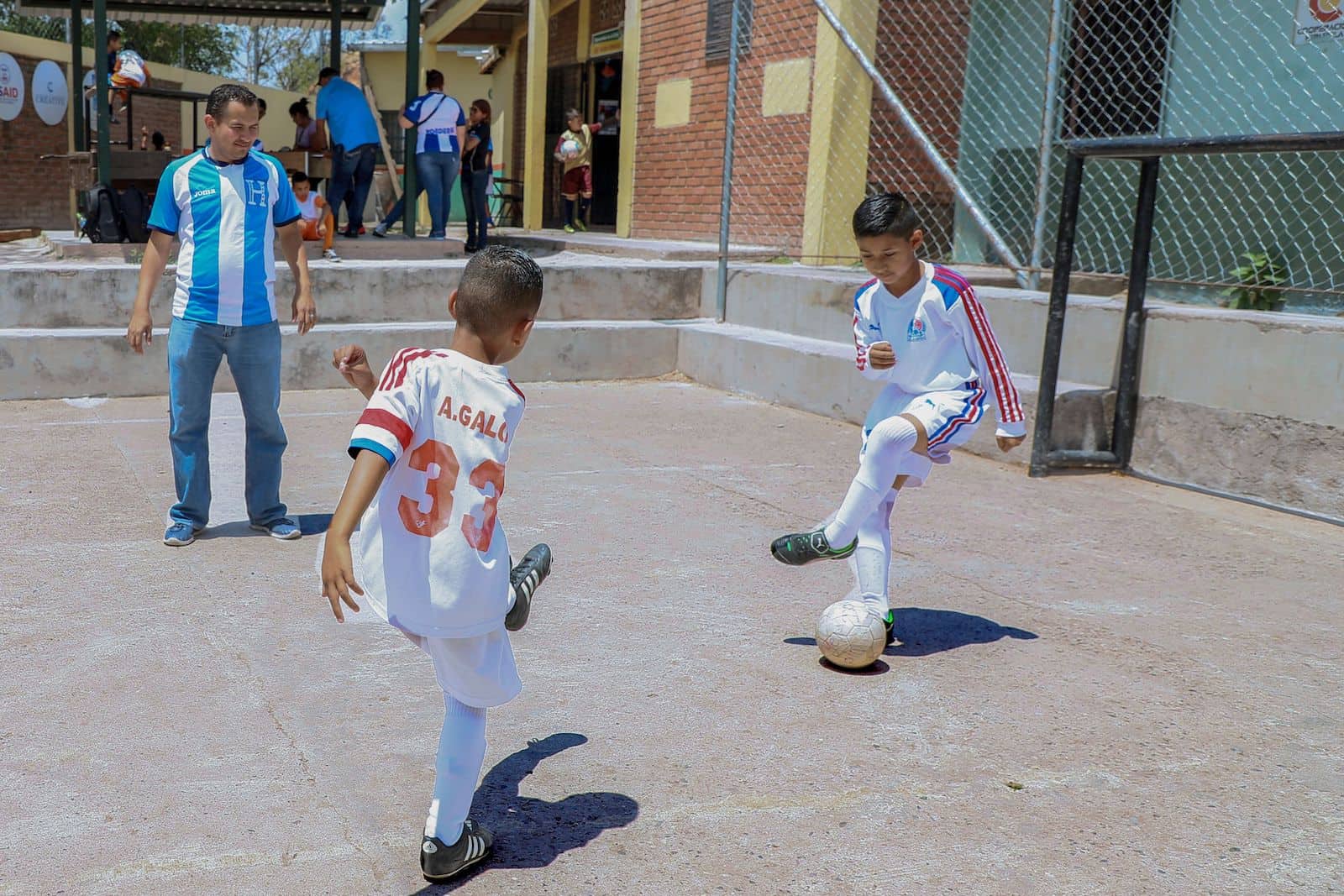 Two boys in white soccer uniforms play soccer in a concrete play yard while a man in a blue and white striped shirt watches.