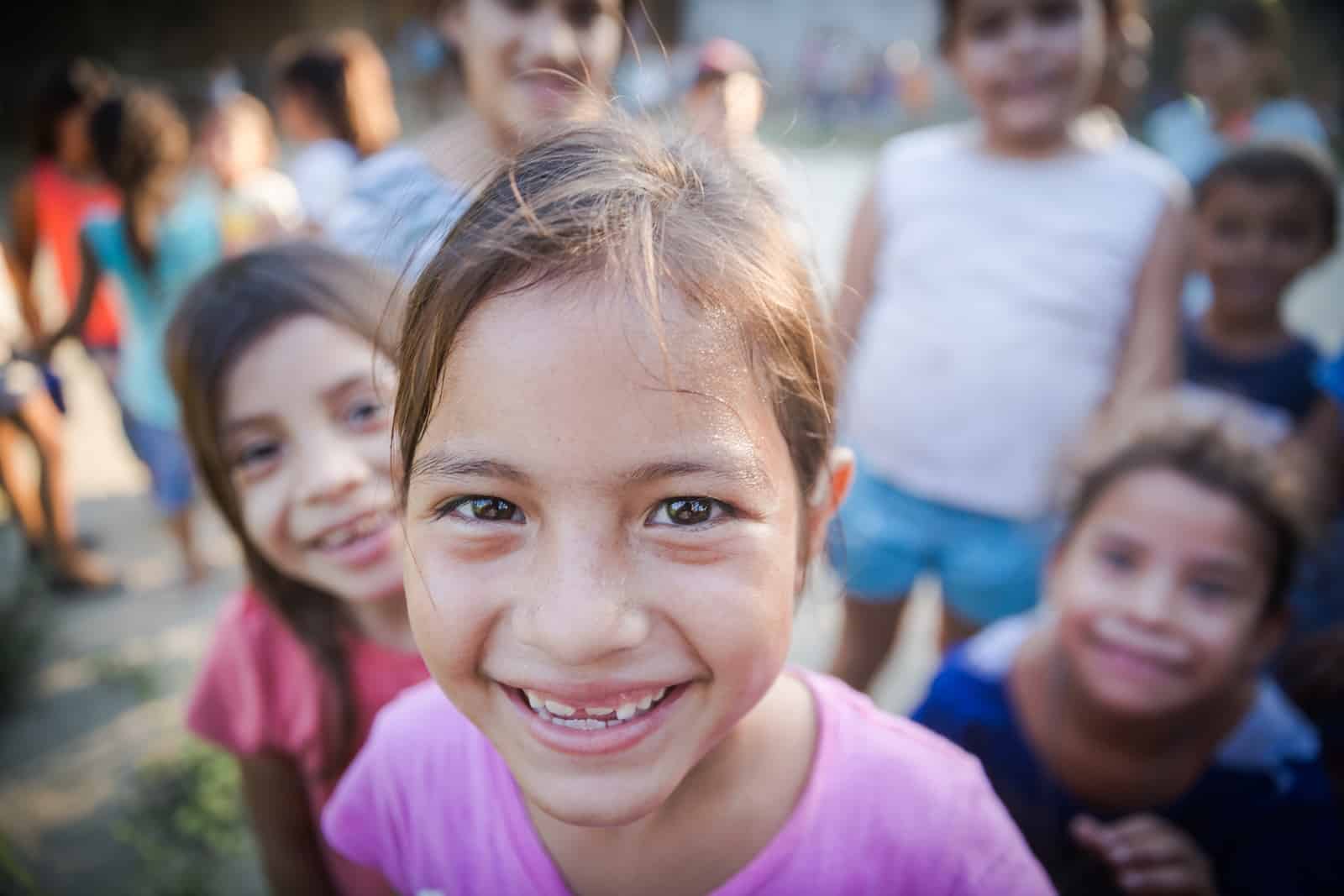 A close-up photo of a girl in a pink shirt and hair in a ponytail smiling at the camera, with several girls behind her smiling.