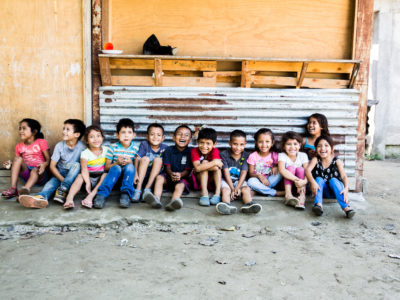 A group of 12 children sit on the ground in a line, smiling. They sit on concrete ground, in front of a corrugated metal sheet and wood wall.