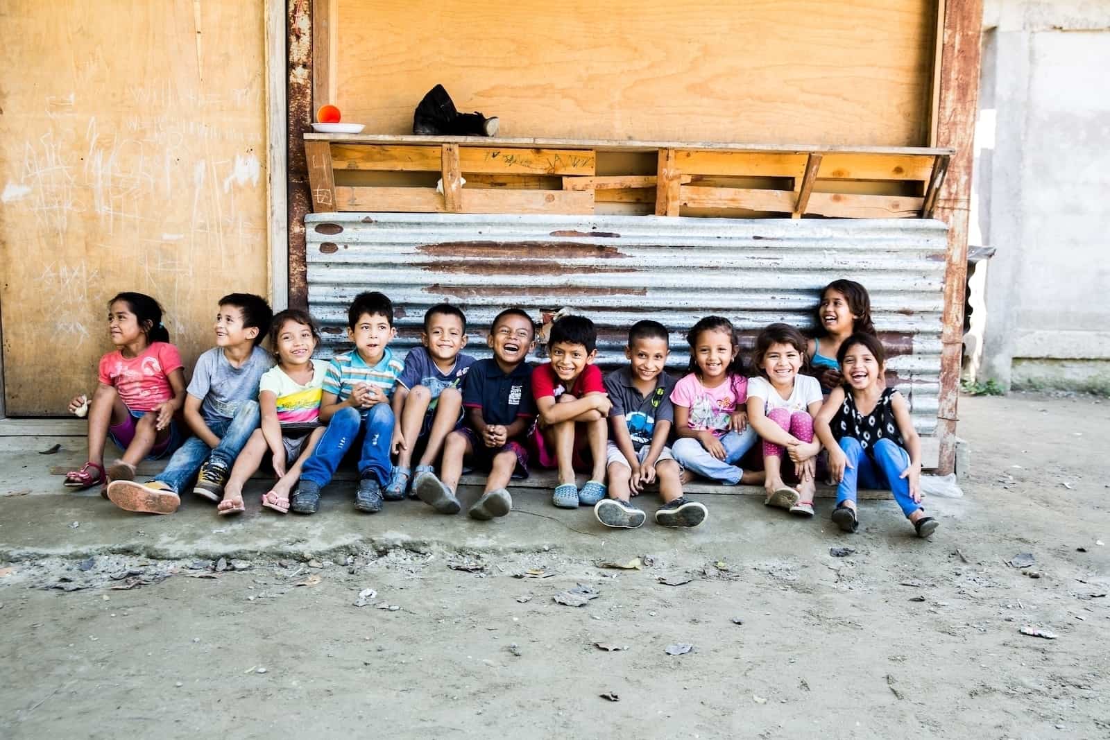 A group of 12 children sit on the ground in a line, smiling. They sit on concrete ground, in front of a corrugated metal sheet and wood wall. 