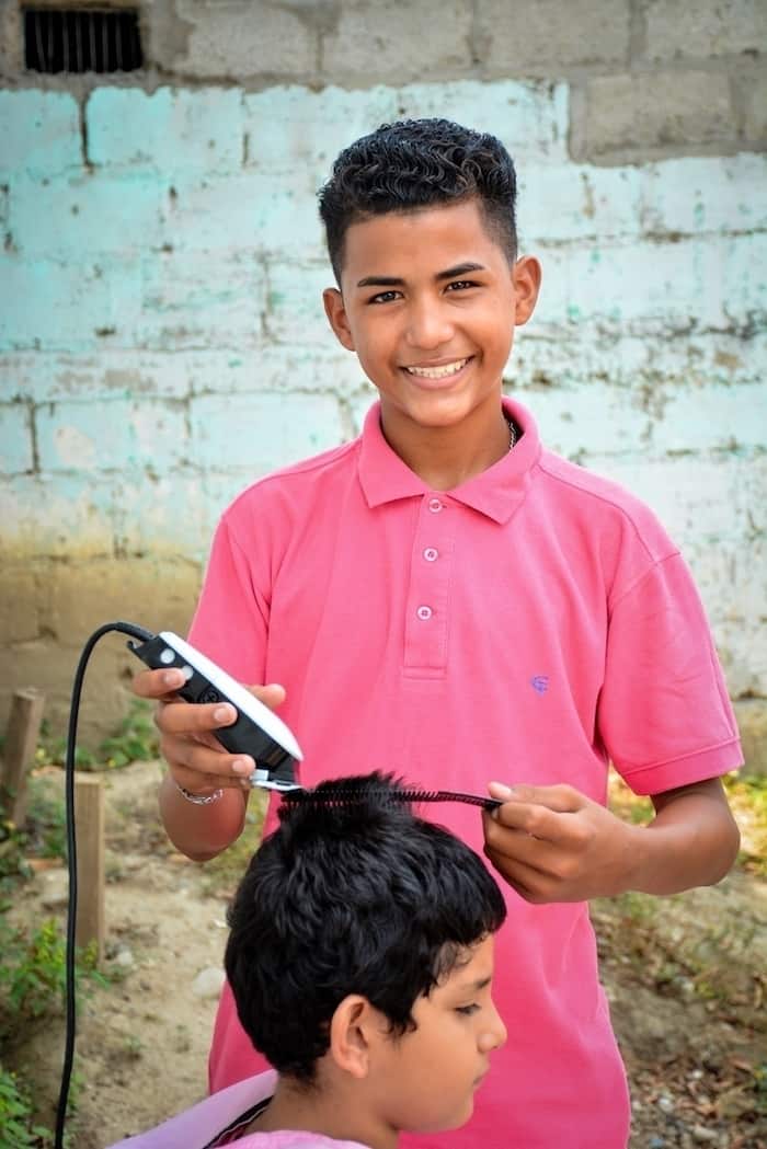 A boy wearing a pink shirt stands, smiling, cutting a boy's hair using clippers and a comb. They are in front of a light green brick wall. 