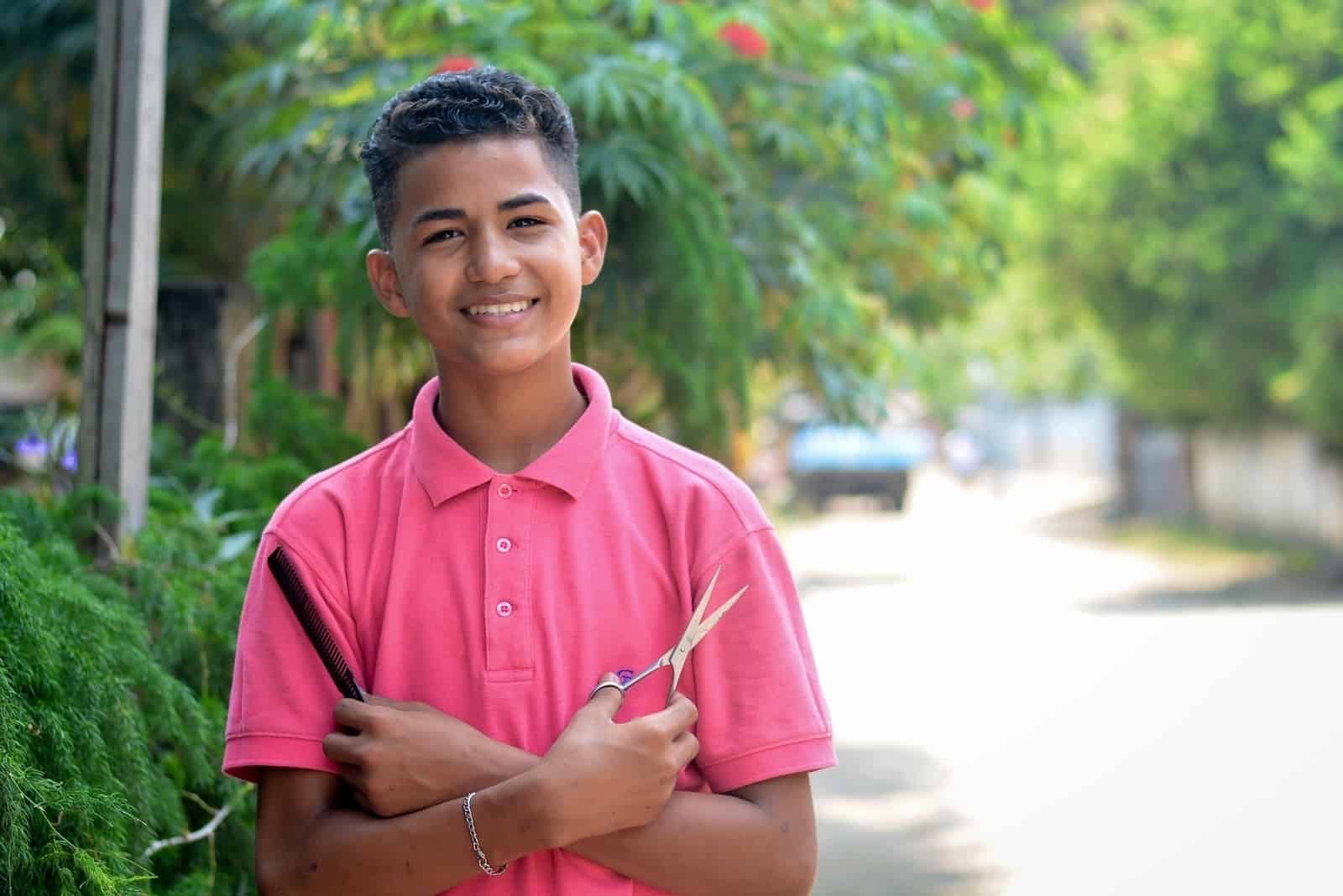 A boy wearing a pink shirt stands, smiling, with his arms crossed holding scissors in one hand and a comb in the other, standing on a street.