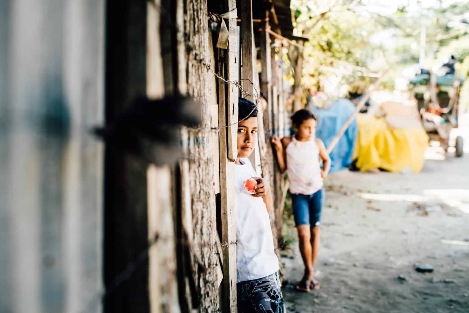A boy stands in a wooden doorway with barbed wire fence wearing a white shirt. In the background is a girl and a dirt road.