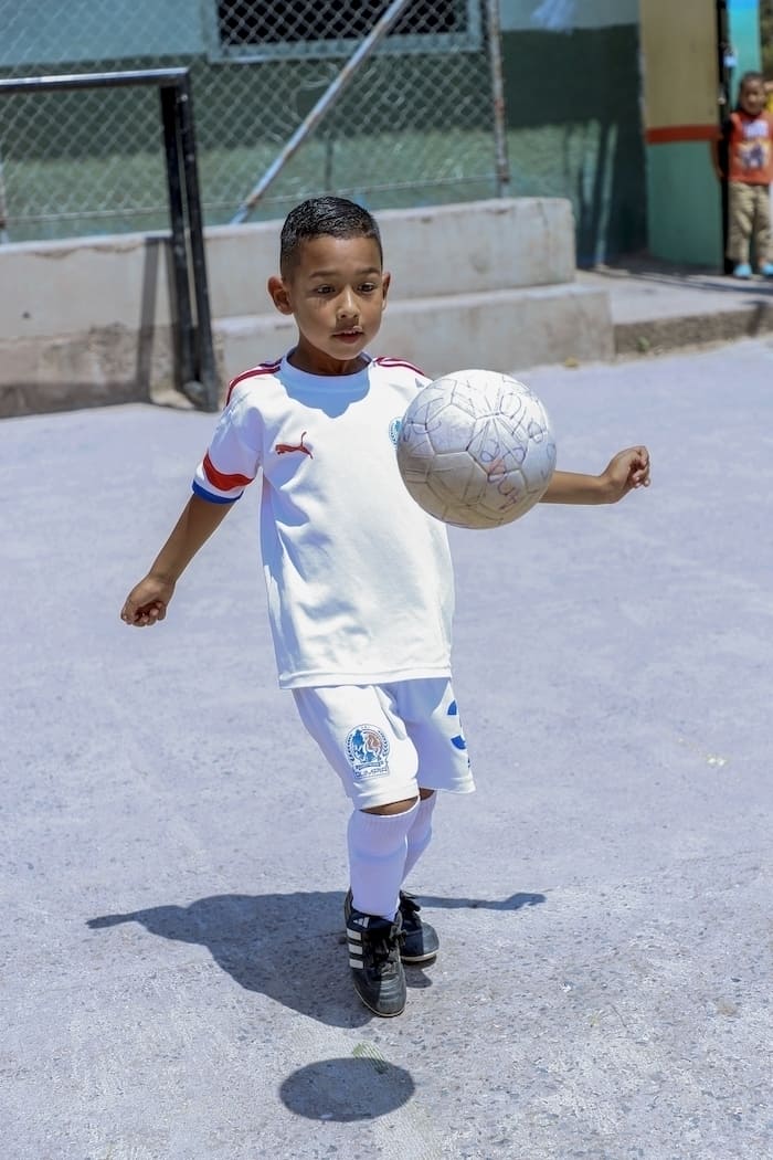 A young boy wearing a white soccer uniform bounces a soccer ball on his knee in a concrete school yard.