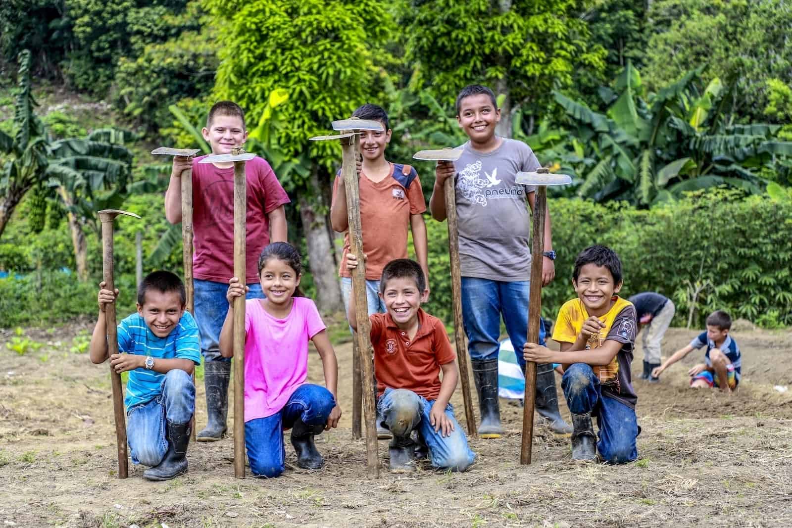 A group of seven children smile at the camera. Three are standing and three are crouching in a field, all holding hoes.