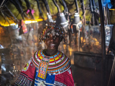 A girl wearing an ornate beaded headdress with silver coins and an ornate beaded necklace stands inside a hut made from wood and leather.