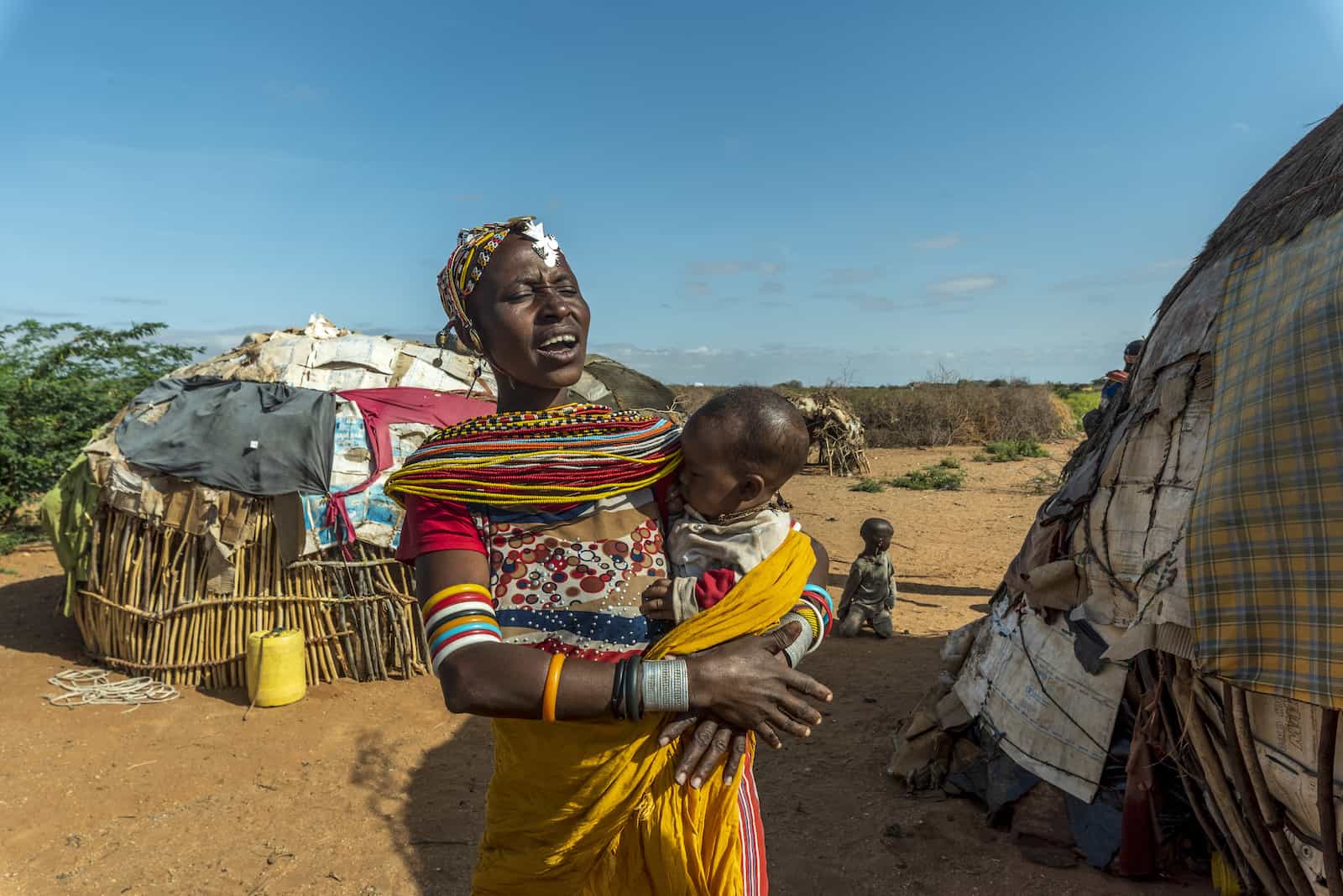 A woman stands outside holding a baby to her chest, standing in front of two small round huts.