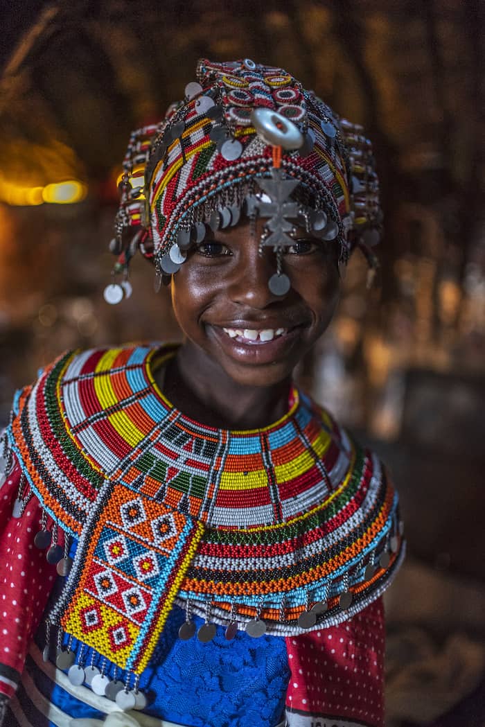 A girl wears an ornate beaded headdress with silver coins and an ornate beaded necklace and smiles at the camera.