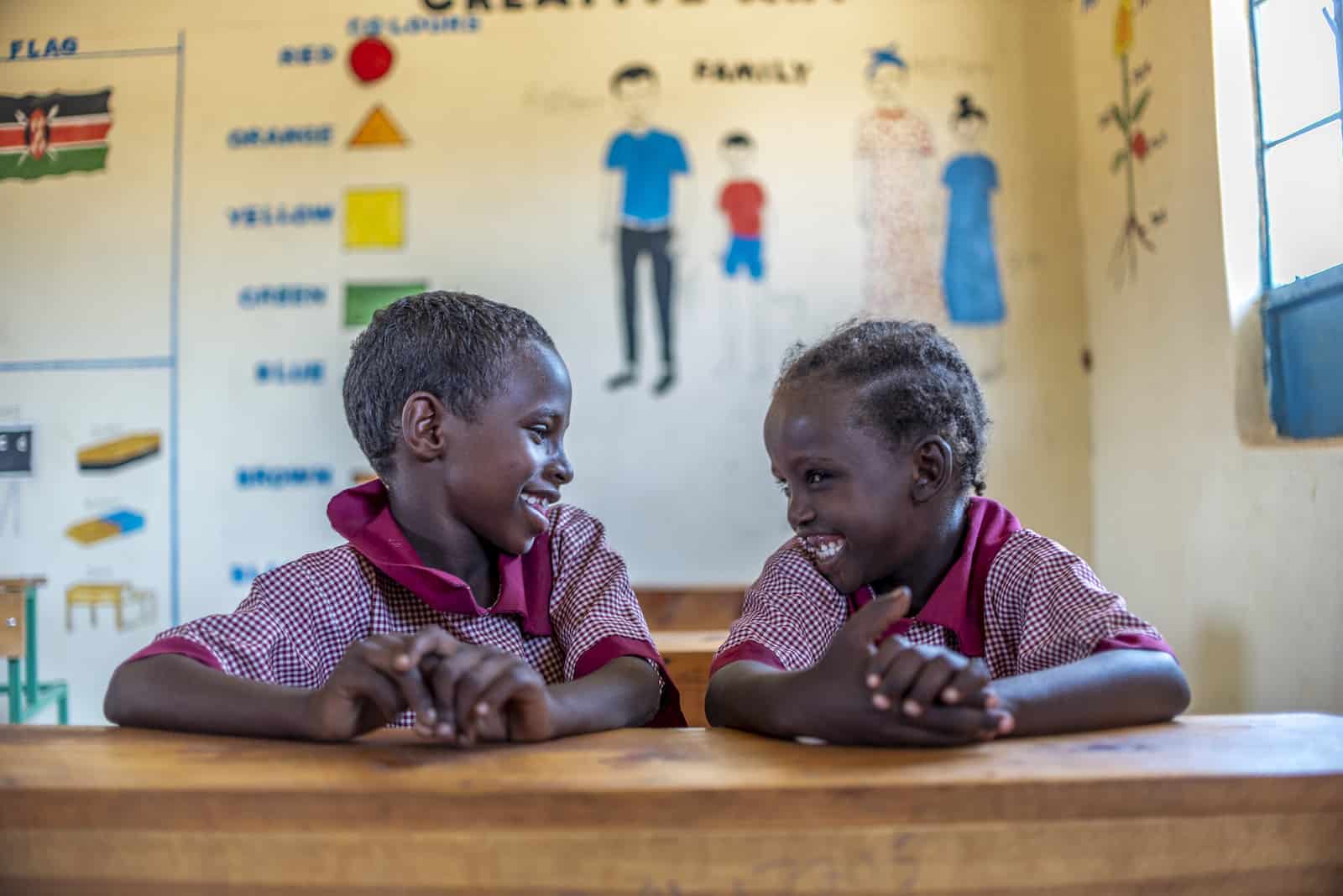 Two young girls where red checked school uniforms sit at a desk in a classroom and smile at each other.