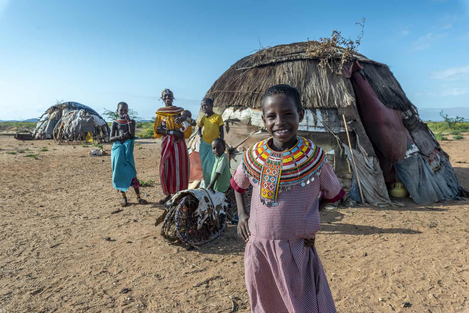 A girl wearing a red school uniform and an ornate beaded necklace stands in front of a rounded hut. Behind her are three children and a woman holding a baby.