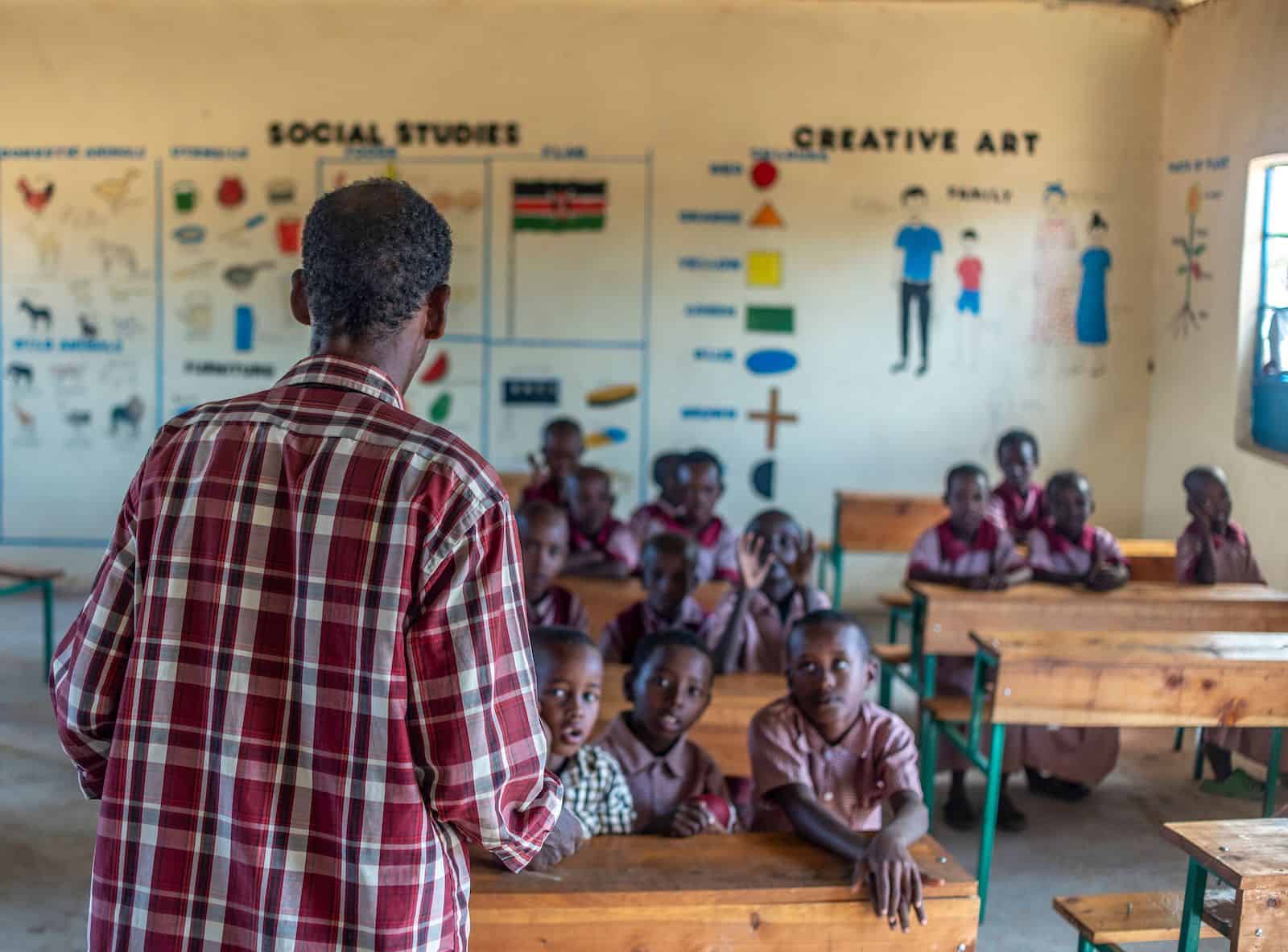 A man in a plaid shirt stands at the front of a classroom, with children wearing red school uniforms sitting at desks and listening.