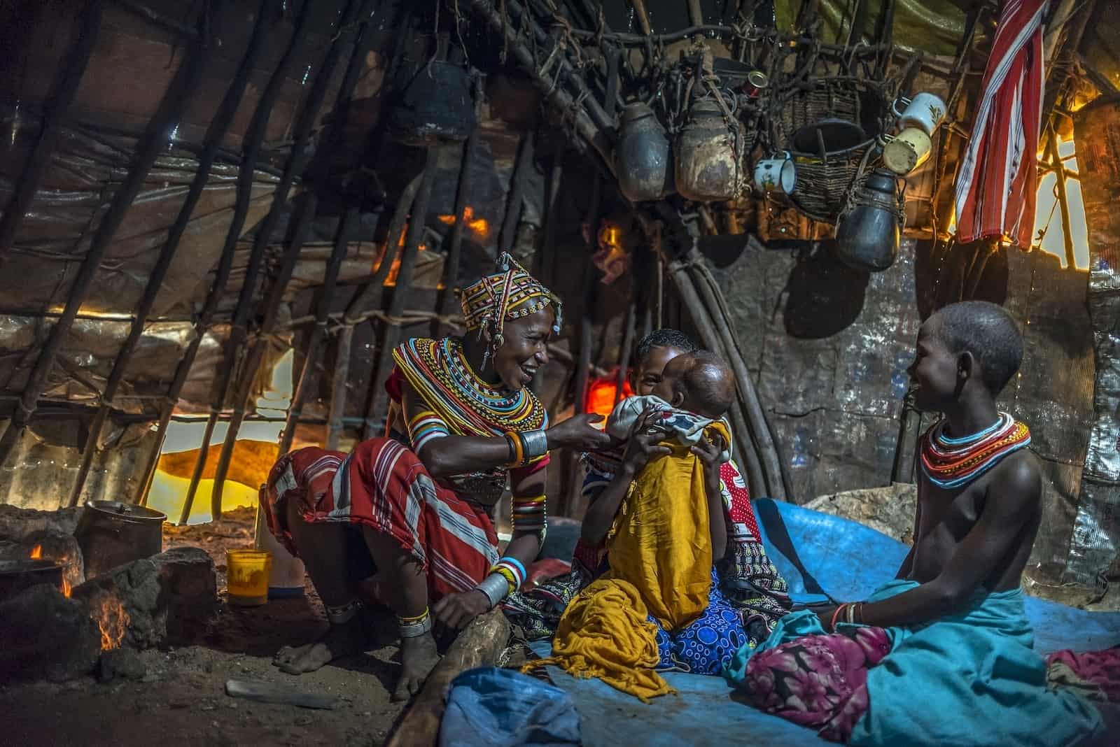 A woman adorned in a beaded headdress and necklace sits on the dirt floor of a small round hut, smiling at a baby being held by a girl sitting next to her. A boy wearing a beaded necklace sits on the other side.