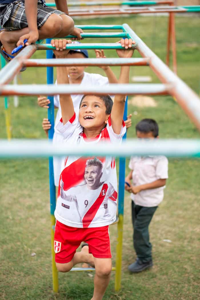 A boy wearing a white shirt and red shorts swings on monkey bars, with several boys behind him waiting in line.