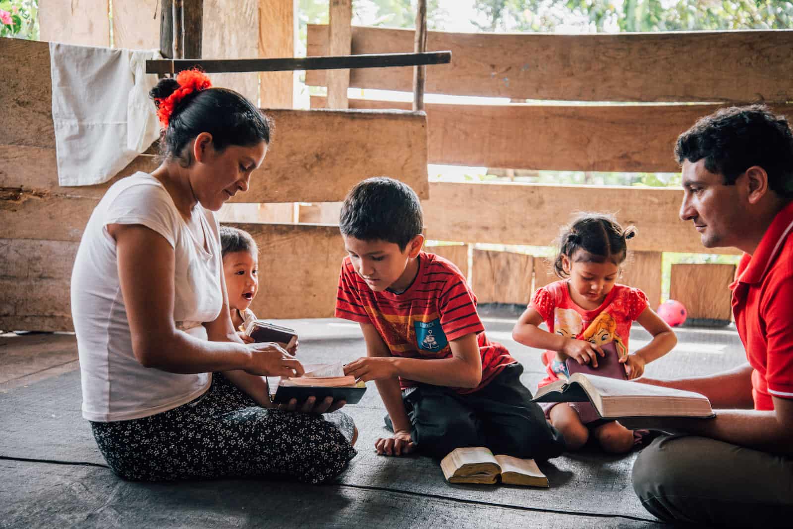A family of five with three young children sit on the floor, each holding a Bible. One boy points to a page in the mother's Bible. They are in a room made from wooden slats. 