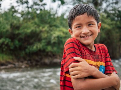 A boy in a red shirt hugs his arms across his chest and smiles. He is standing in front of a river in the Amazon jungle.