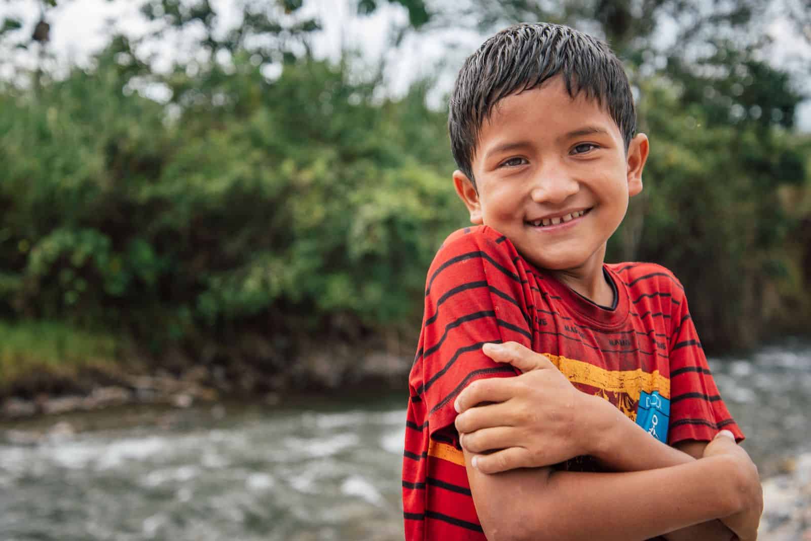 A boy in a red shirt hugs his arms across his chest and smiles. He is standing in front of a river in the Amazon jungle. 
