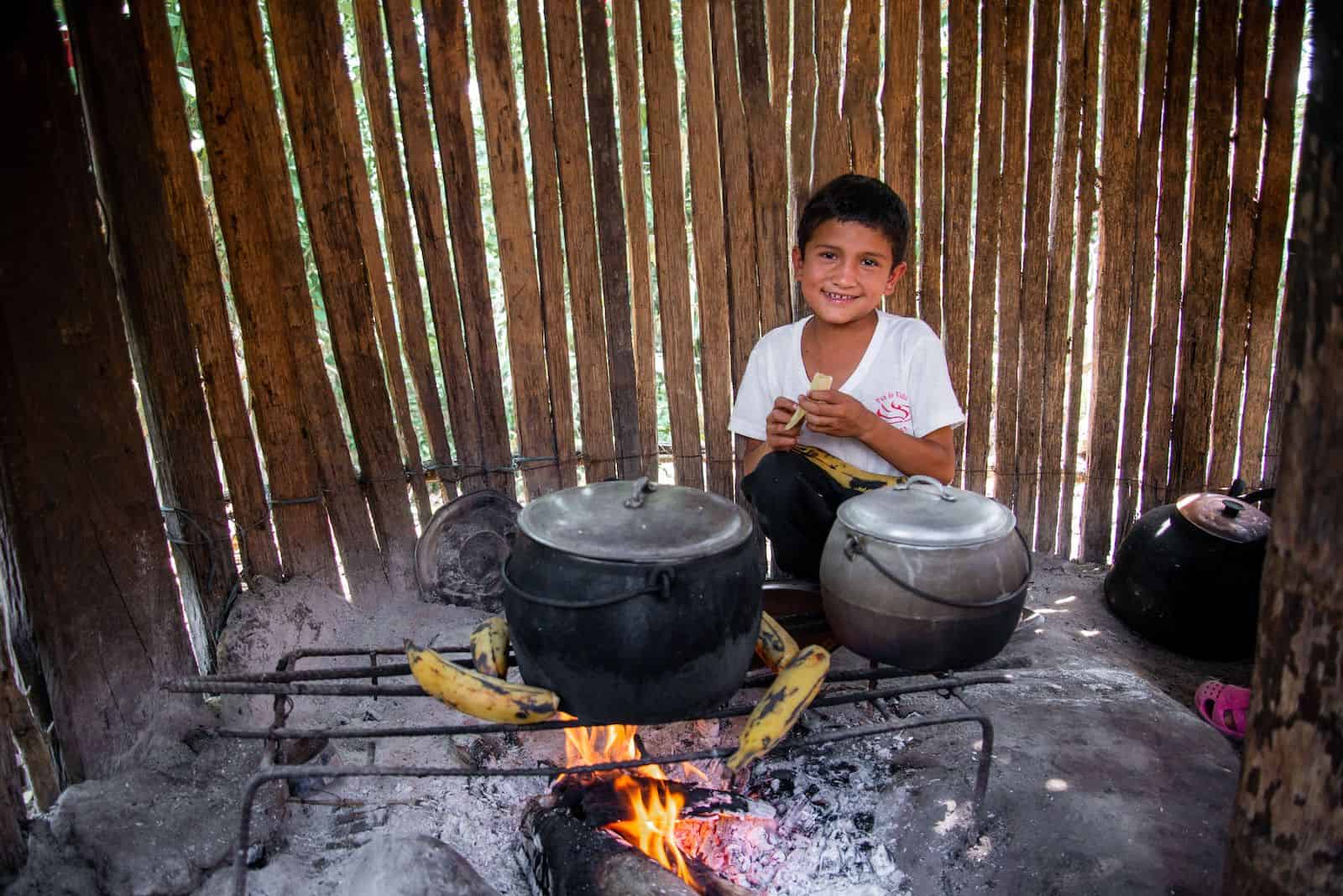 A boy wearing a white shirt crouches in front of a wood fire that has a wire frame over it with several plantains on it and two black pots. 