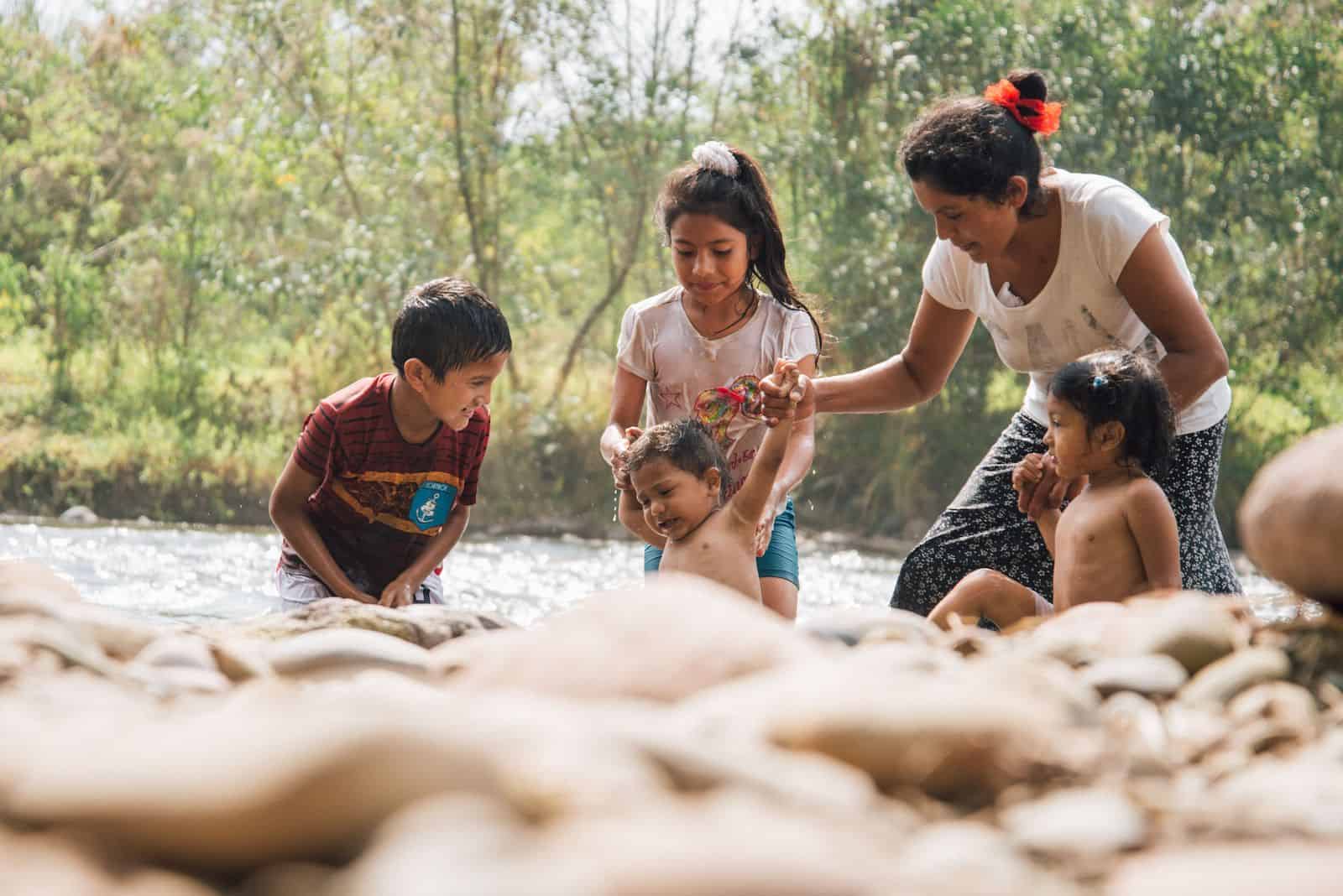 A mother and four children stand in a river surrounded by green foliage. A toddler is being held by the hands by the mother and sister. 