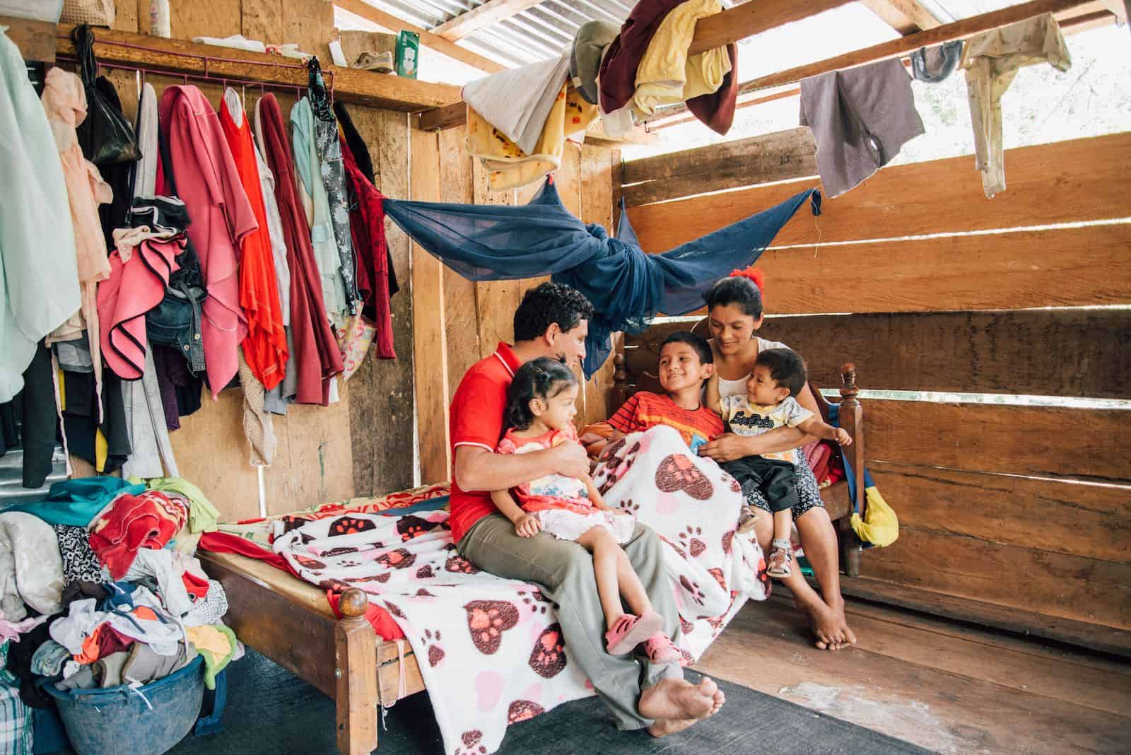 A family of five sits on a bed in a room made from wood and a iron sheet roof. Sunlight comes in through cracks of the wood, and clothing hangs on the walls. The dad holds a young girl on his lap and the mom holds a young boy on his lap, all looking at and smiling at a boy under the covers.