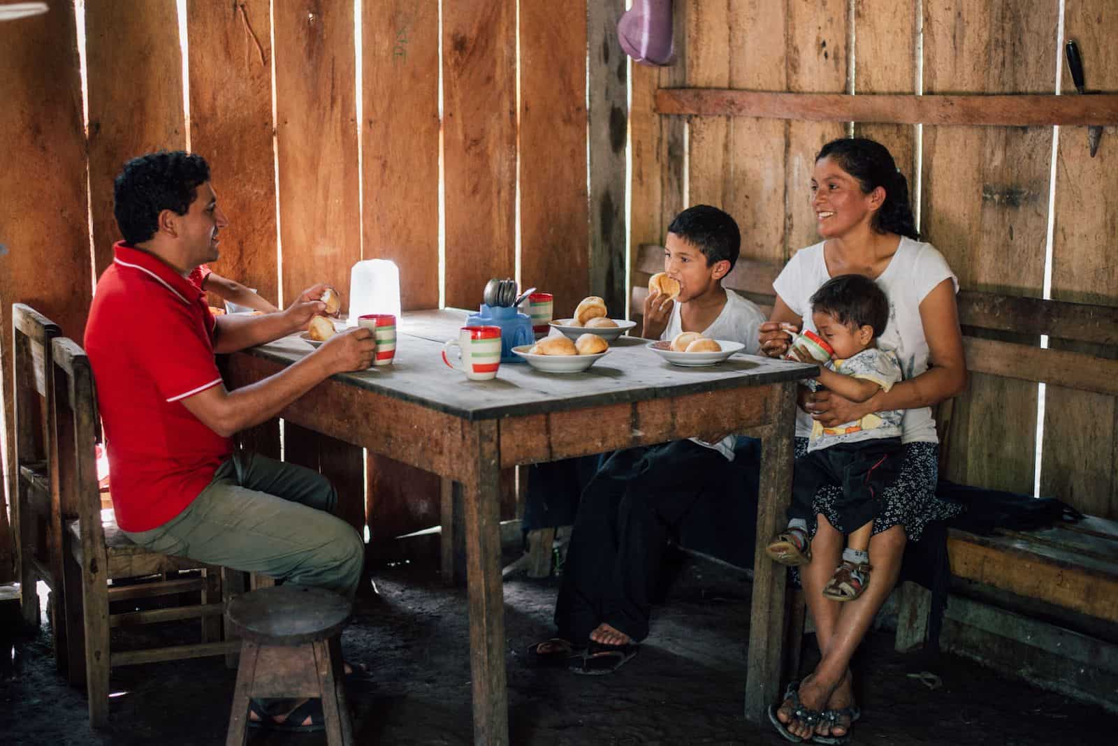 A family of five sits at a wooden table with mugs and bowls with rolls in them. The room is made of wooden slats. 