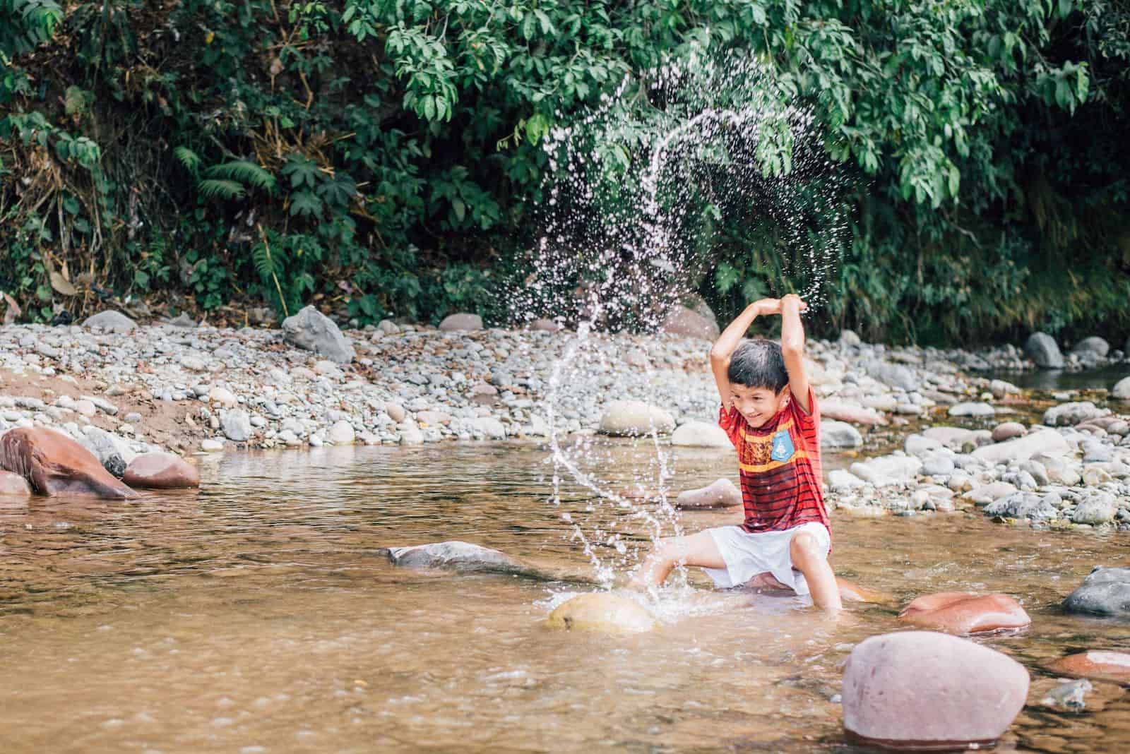 A boy wearing a red shirt and white shorts sits in a shallow riverbed surrounded by green foliage, and splashes water. 