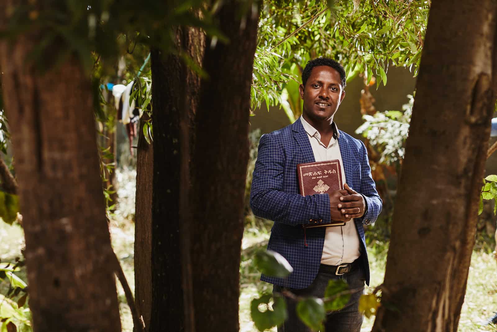 A man stands in a group of trees wearing a suit, holding a Bible and smiling into the camera.