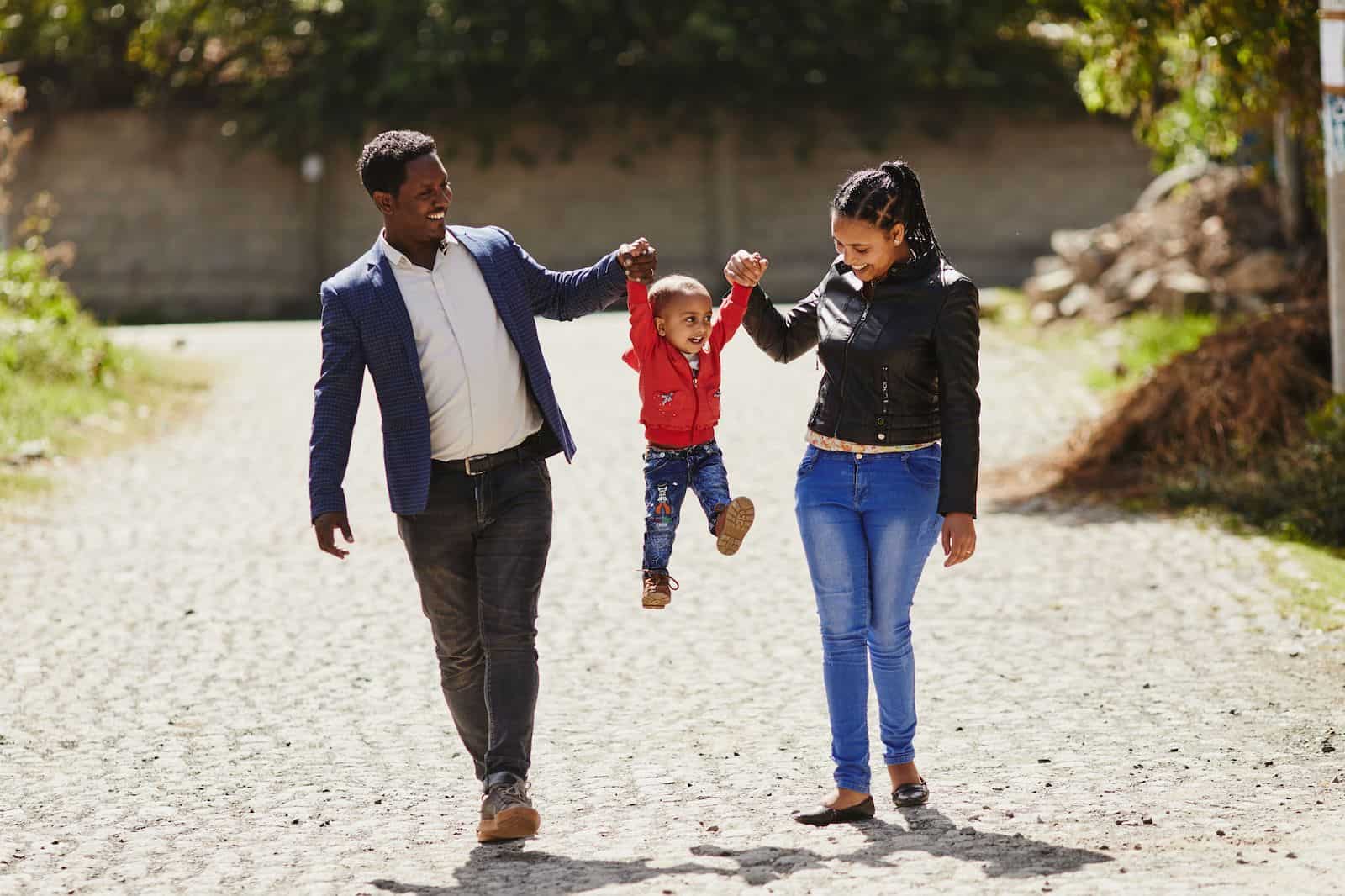 A man wearing a suit and a woman wearing jeans and a black jacket hold the hands of a toddler and swing him in the air, while walking down a cobblestone street.