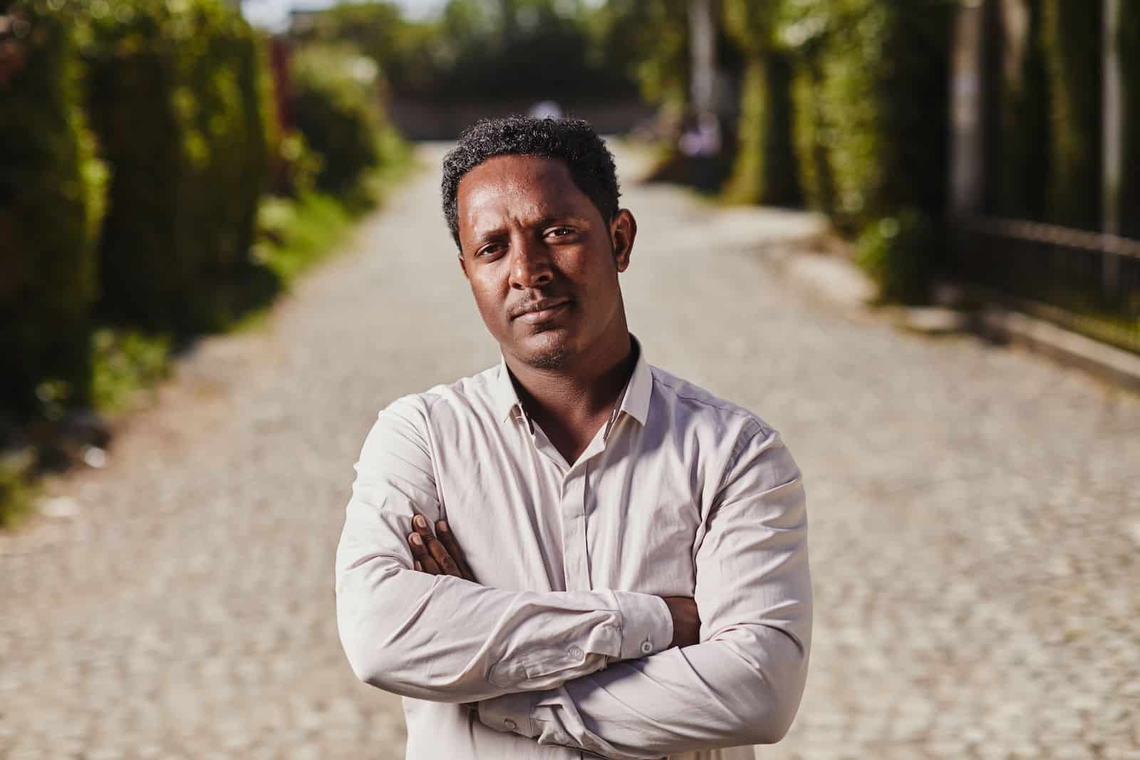 A man stands with his arms crossed in front of him, wearing a white button-up shirt, looking solemnly into the camera. He is standing on a cobblestone street surrounded by green trees. 