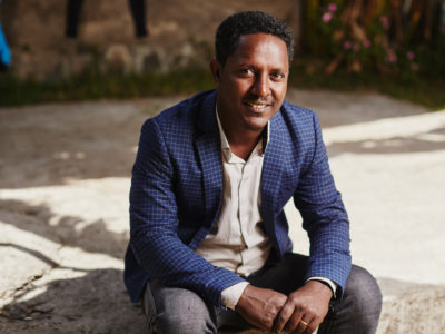 A man wearing a blue suit jacket, white shirt and gray pants, sits on a concrete step with his hands clasped, smiling at the camera.