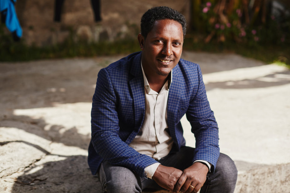 A man wearing a blue suit jacket, white shirt and gray pants, sits on a concrete step with his hands clasped, smiling at the camera.