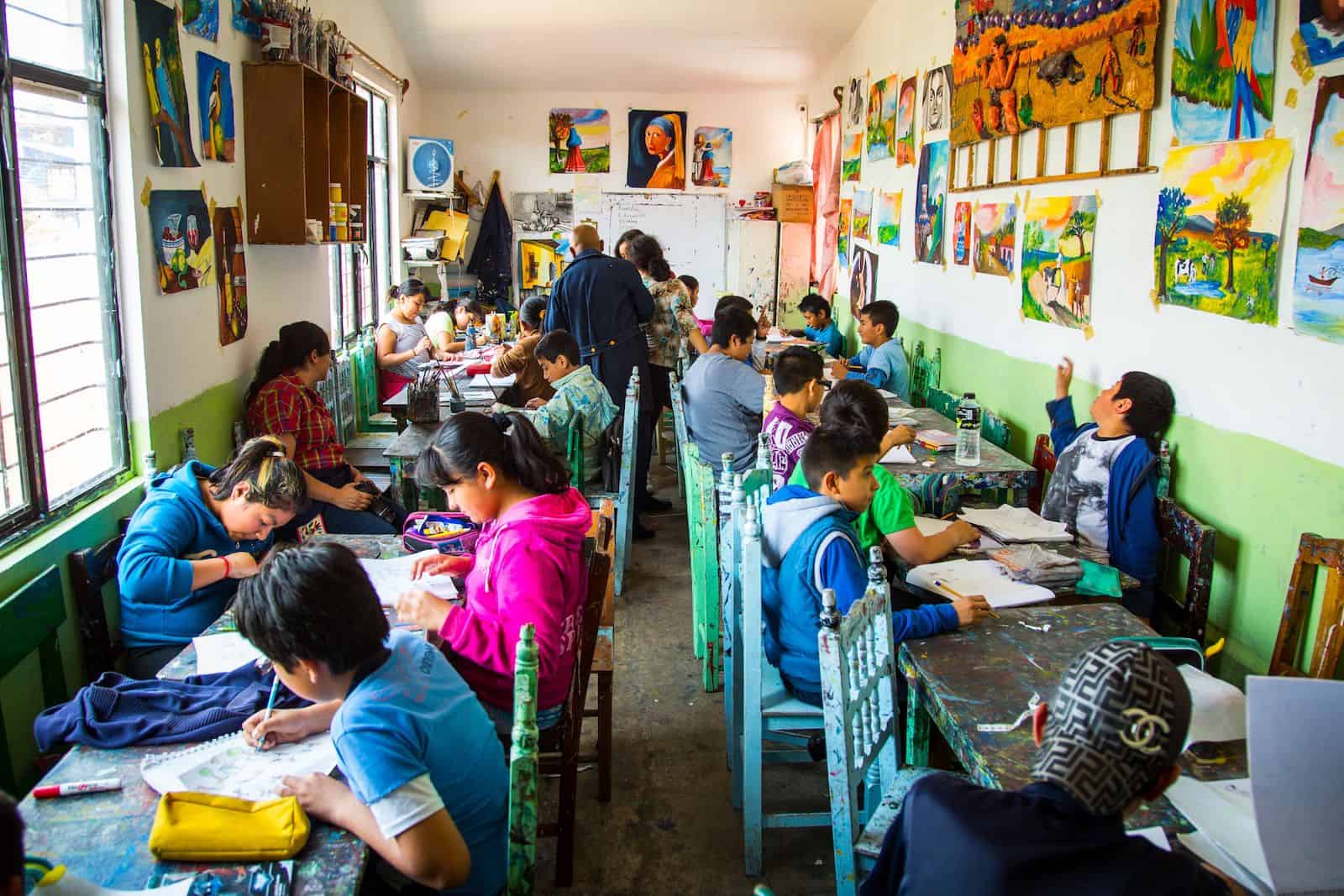 In a classroom full of brightly colored chairs and tables, children paint on white paper. The walls of the classroom are covered in paintings.