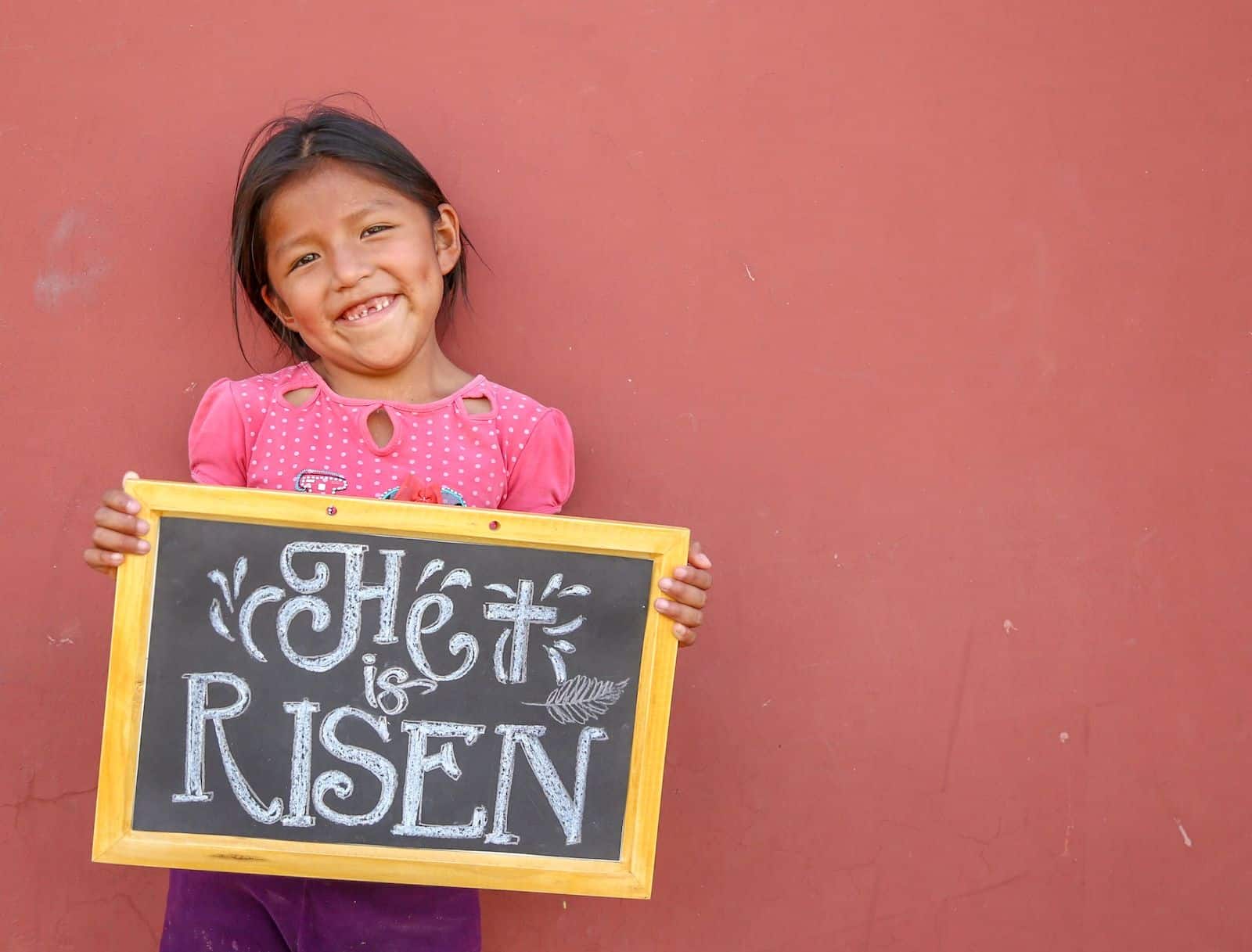A girl missing her front tooth smiles, holding a chalkboard that says, "He is risen." She is wearing a pink shirt and purple pants and stands in front of a coral colored wall.
