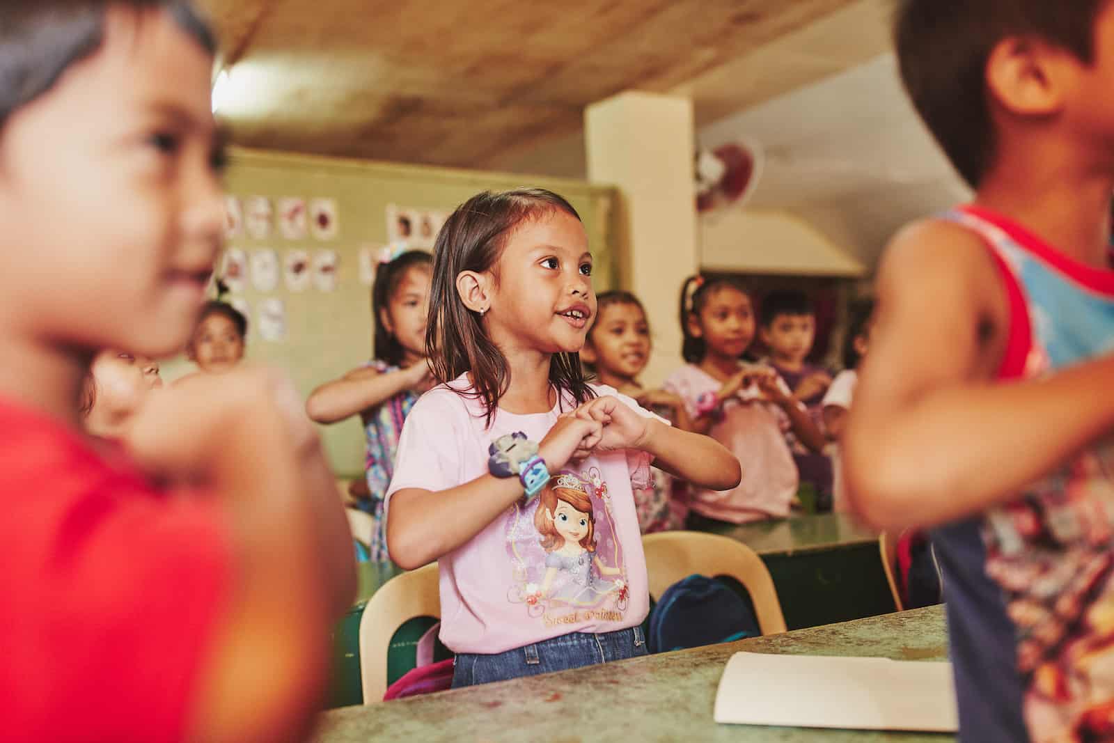 A young girl in the Philippines in a pink shirt makes a heart shape with her hands in front of her, singing and looking to the front of the class. She is in a classroom surrounding by kids doing the same thing. 