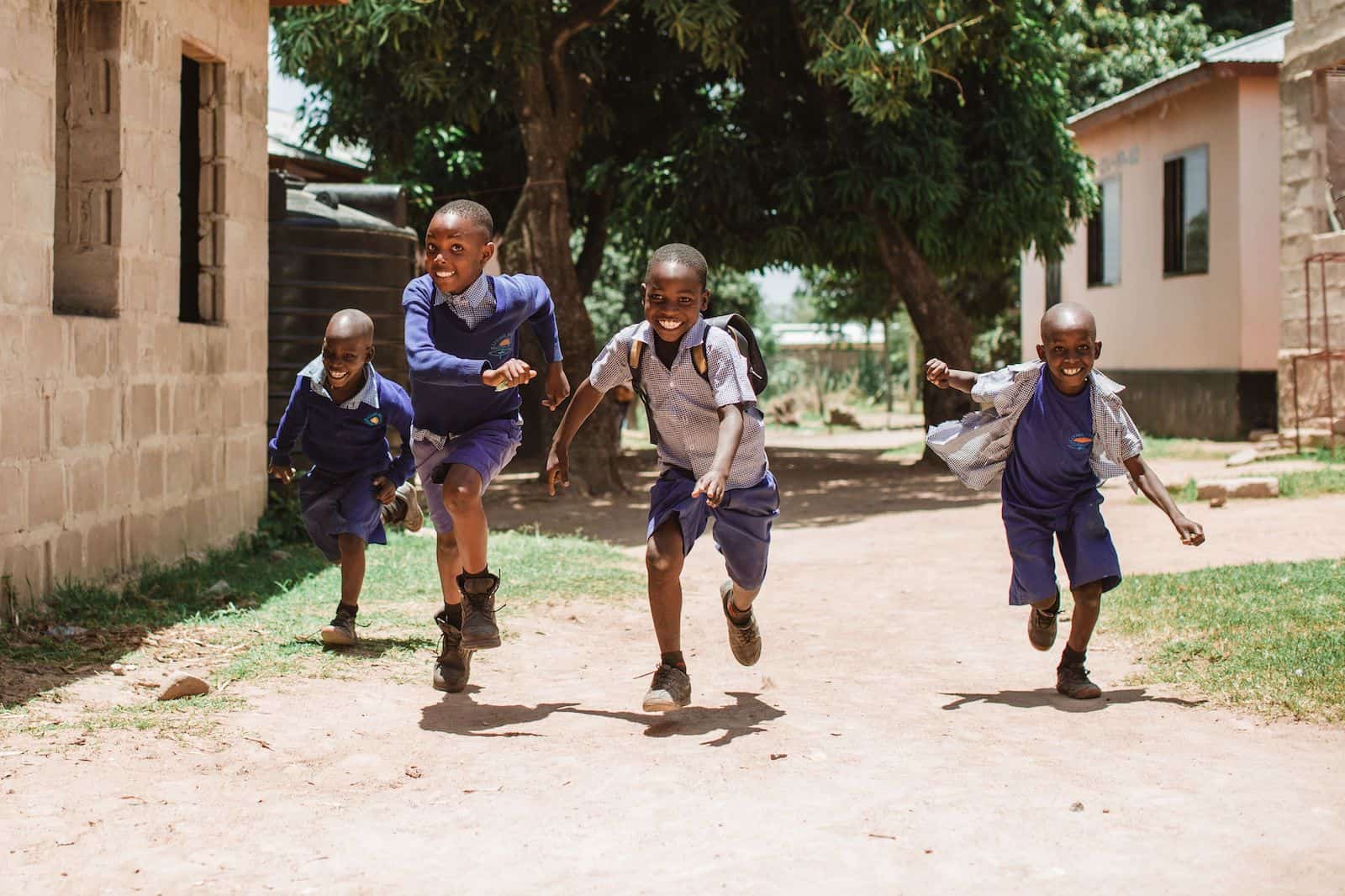 Four boys in Tanzania wearing purple school uniforms run towards the camera, smiling, down a dirt road.