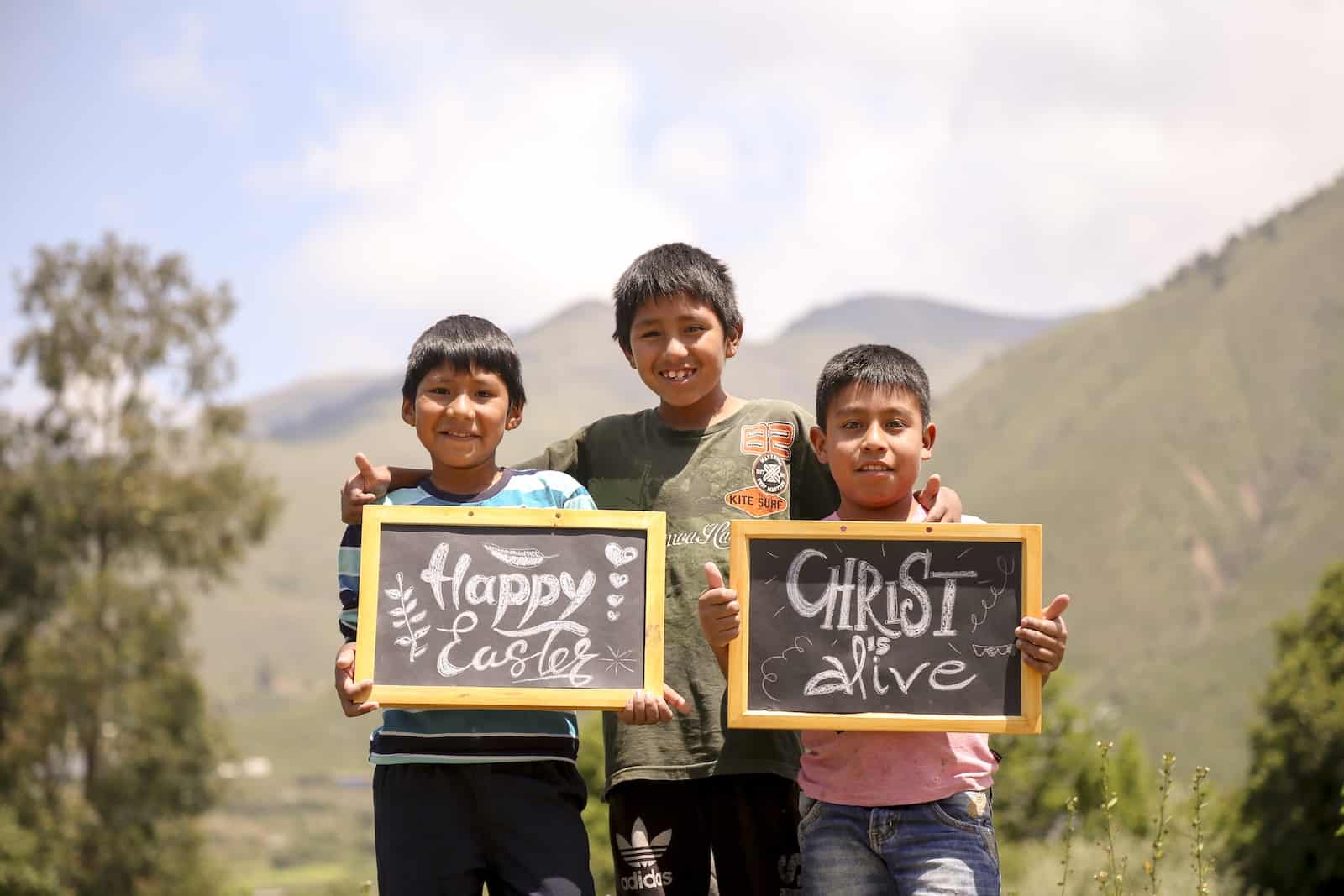 Three boys stand in front of green hills in Bolivia. The one on the left holds a sign that says, Happy Easter. The one on the right holds a sign that says, Christ is alive.