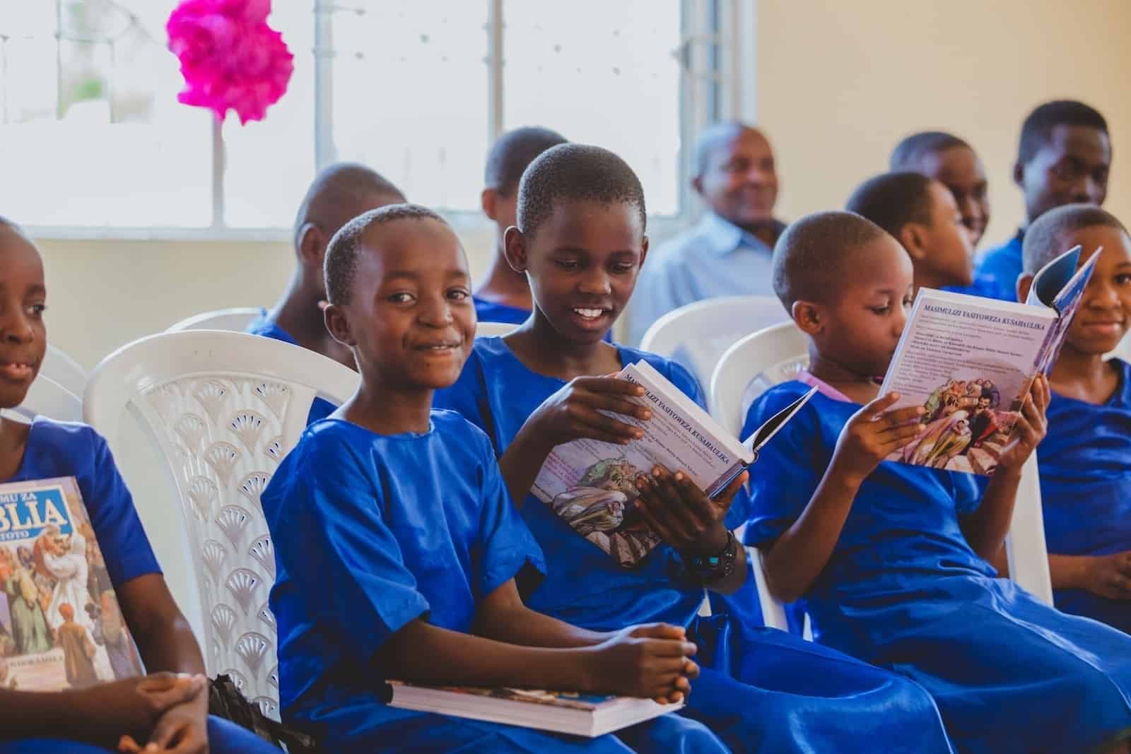 A group of girls in Tanzania wearing blue uniform dresses sit on white plastic chairs, each holding and reading Bibles. The girl in the foreground smiles at the camera.