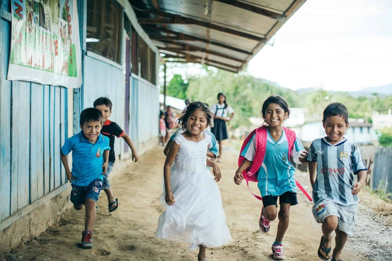 A group of children run down a path in Peru, next to a row of blue classrooms.