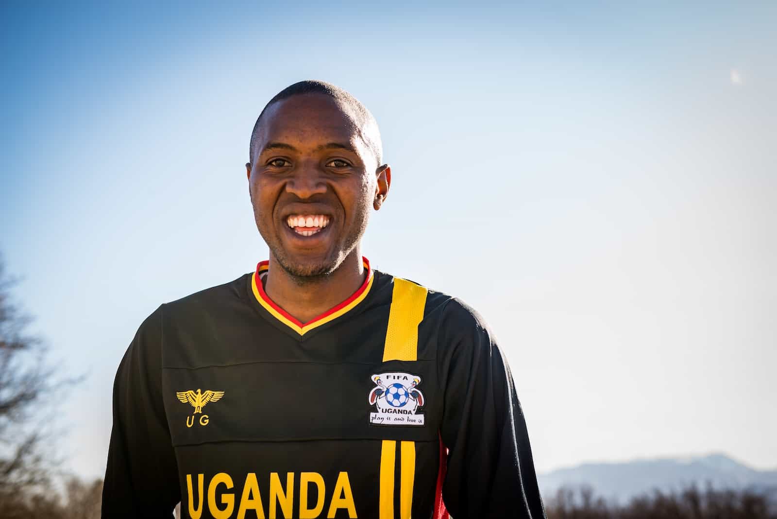 A man smiles, wearing a black shirt that says, "Uganda."