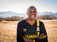 A man wearing a black jersey that says Uganda crosses his arms and smiles. He stands outside with trees and mountains in the distance.